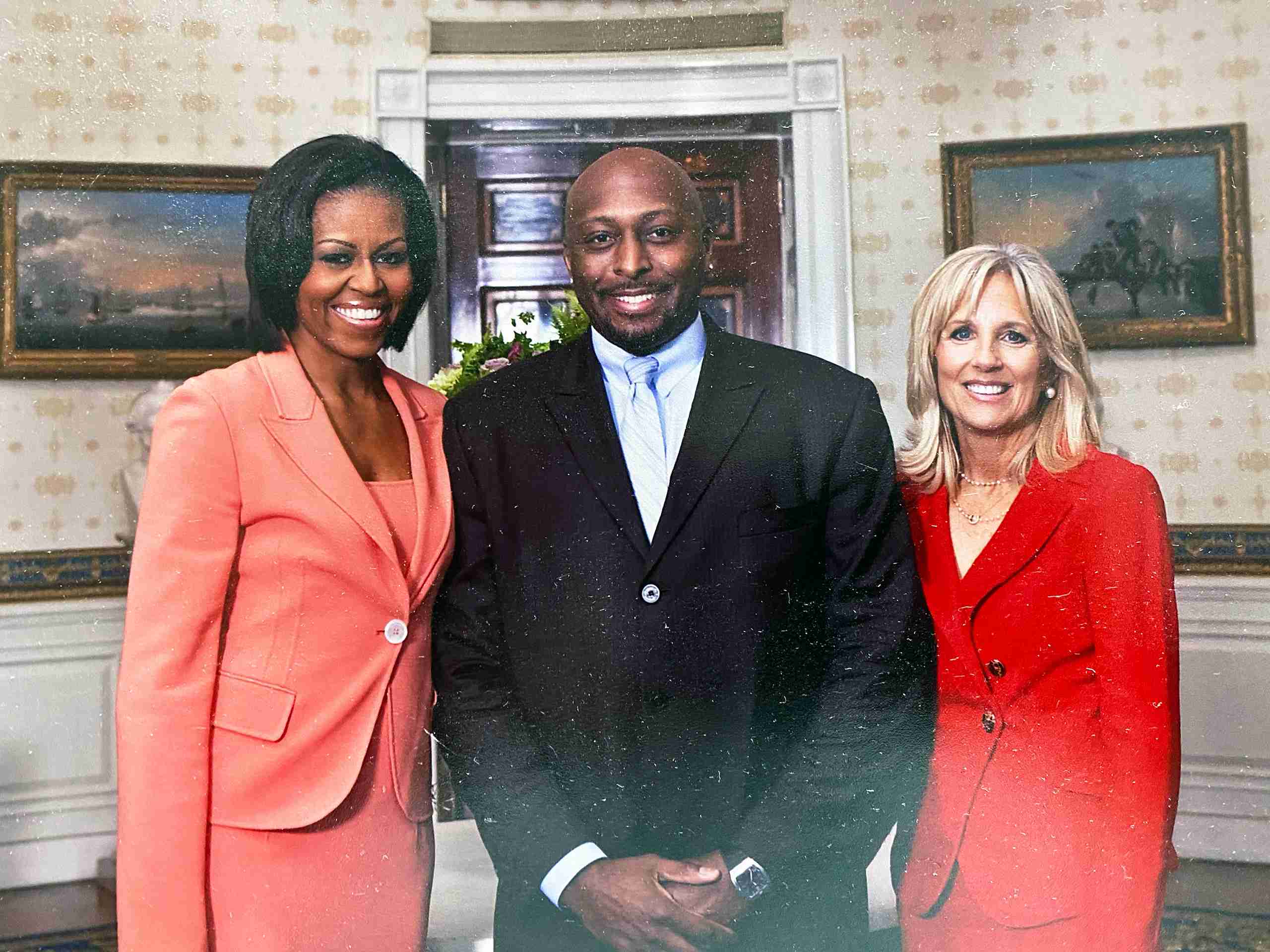 Three people in formal attire stand together in a decorated room with framed paintings and a floral arrangement in the background.
