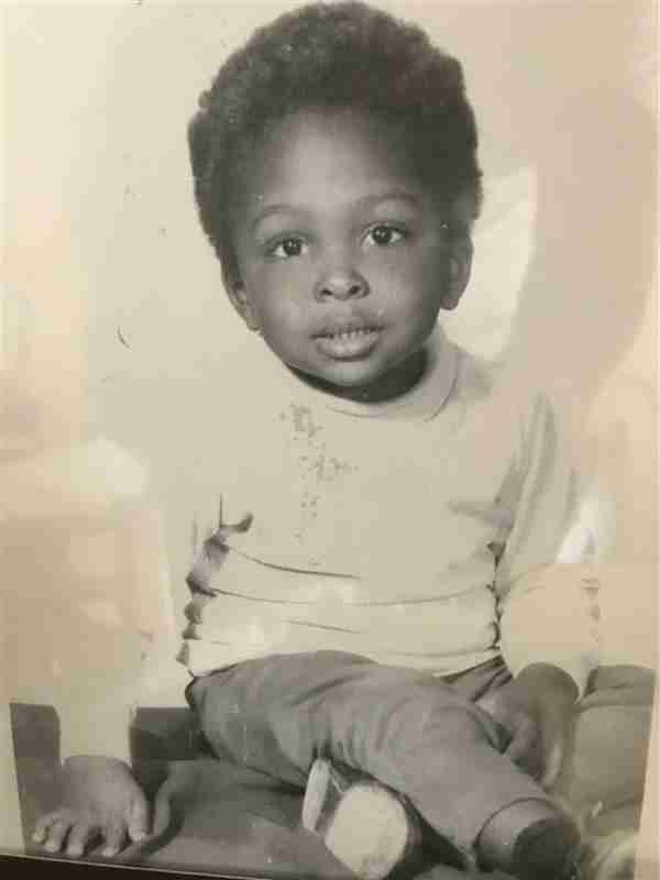 Black and white photo of a young child with short curly hair, wearing a light-colored sweater and pants, sitting and looking toward the camera.