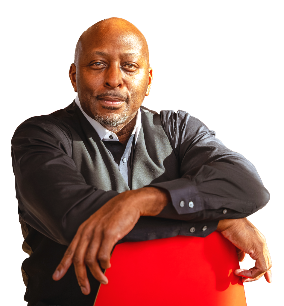 A man in a black shirt and vest sits on a red chair, looking at the camera with a neutral expression against a plain white background.