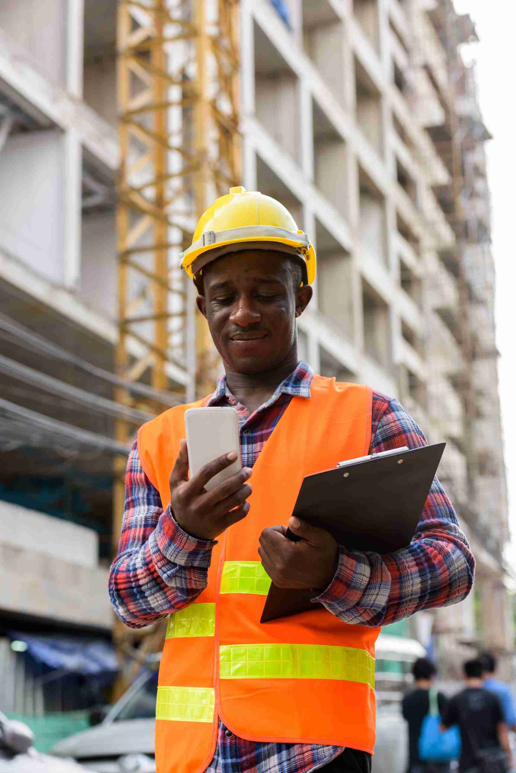A construction worker in a safety vest and helmet holds a clipboard and checks a smartphone at a building site, possibly participating in a construction pre-apprenticeship program.