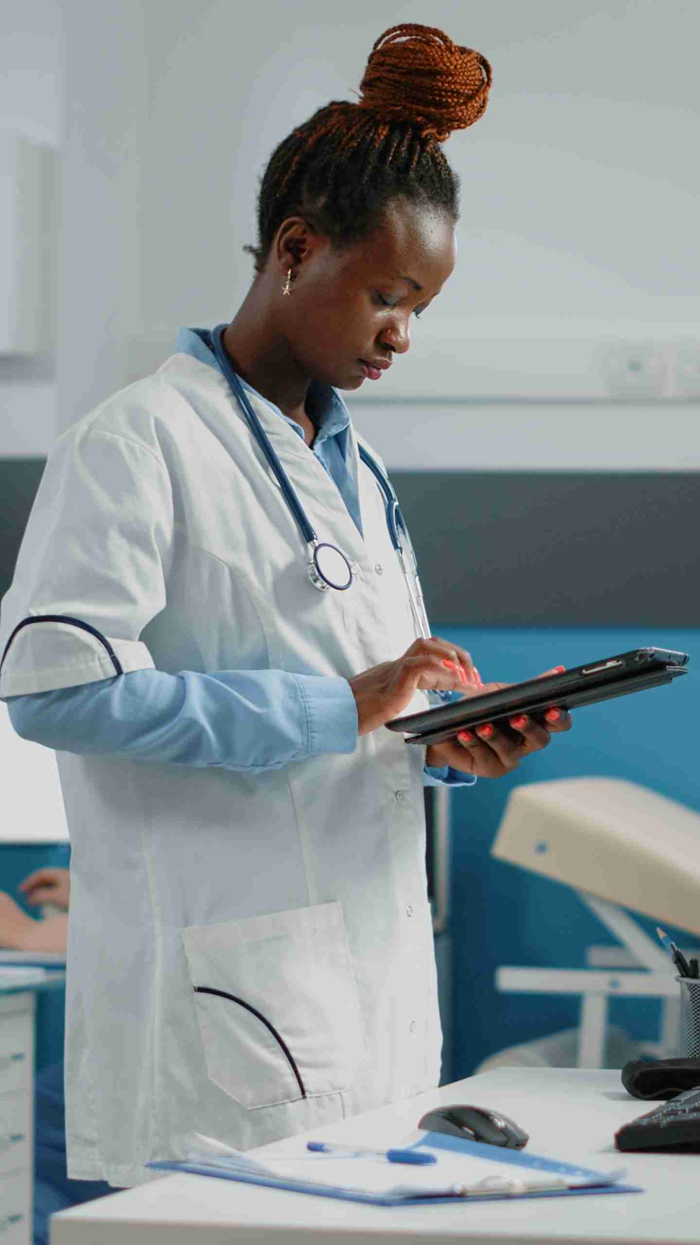 A doctor in a white coat and stethoscope uses a tablet in a medical office, collaborating with the Community Health Worker role to enhance patient care.