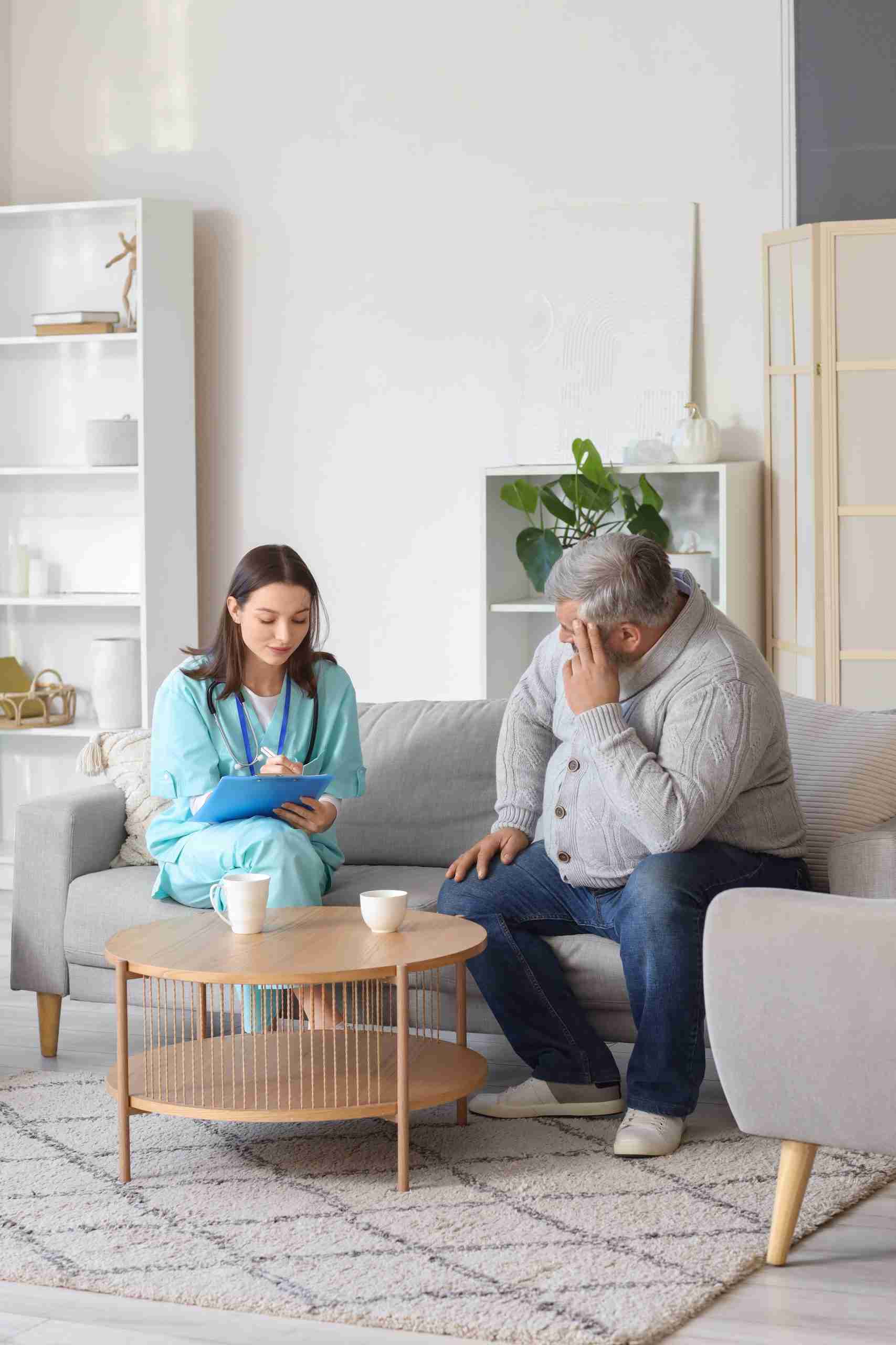 A woman in scrubs, fulfilling a Community Health Worker role, talks to an older man on a living room couch, holding a clipboard and pen. Two cups rest on the coffee table in front of them.
