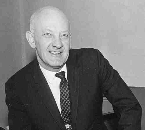 A middle-aged man in a suit and tie sits on a chair, smiling at the camera with his hands resting on his knees, posing confidently for a press kit.