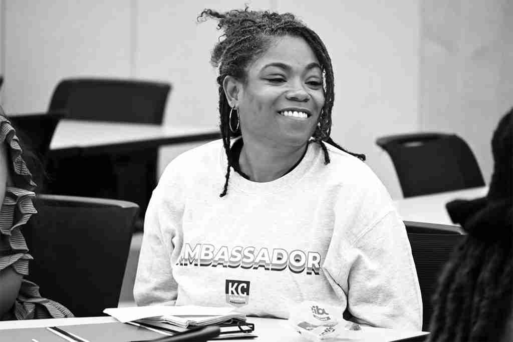 A woman wearing a sweatshirt labeled "AMBASSADOR" sits at a table in a classroom, smiling, as she prepares materials for a press kit.