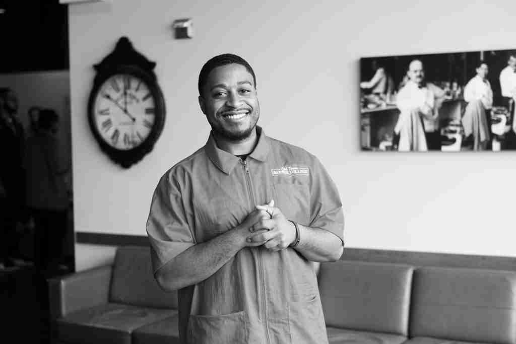 A man smiles while standing in front of a couch, with a wall clock and a black-and-white photo in the background—perfect for any press kit.