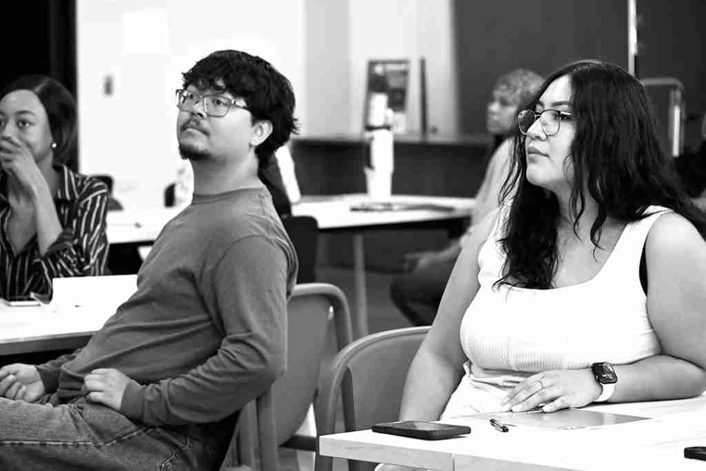 A group of people sit at desks in a classroom, attentively listening to something out of view, perhaps reviewing materials from a press kit.