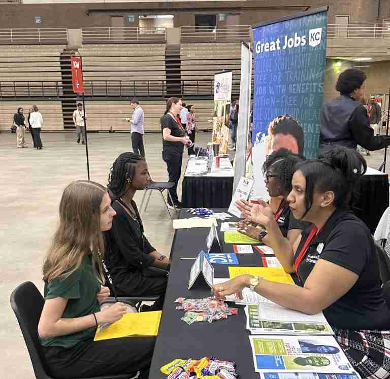 Two young women sit across from two recruiters at a booth during a job fair in a large indoor venue, with informational brochures and candy on the table.