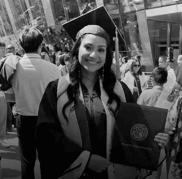A young woman in a graduation cap and gown smiles while holding a diploma folder among a crowd outdoors, perfect for inclusion in any press kit celebrating academic achievement.