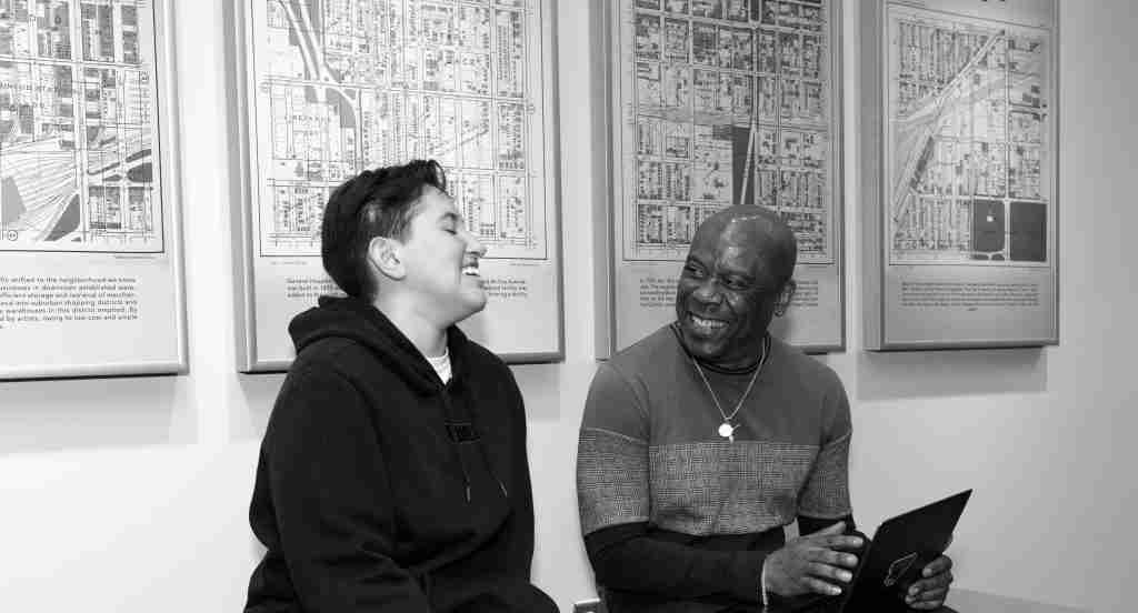 Two people sit and smile at each other in front of maps on the wall; one holds a tablet, possibly discussing materials for a press kit, and both appear engaged in conversation.