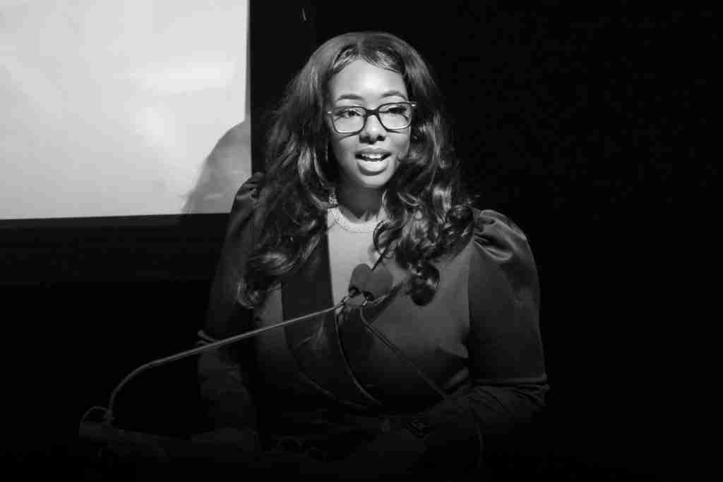 A woman with long hair and glasses speaks at a podium, illuminated by a spotlight, with a press kit and a screen partially visible in the background.