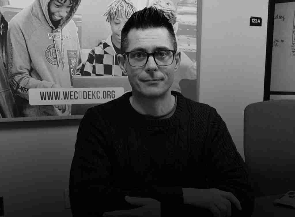 A man with glasses sits at a desk in an office; a press kit rests nearby, while a poster of three people and a visible "wecodekc.org" sign hang on the wall behind him.