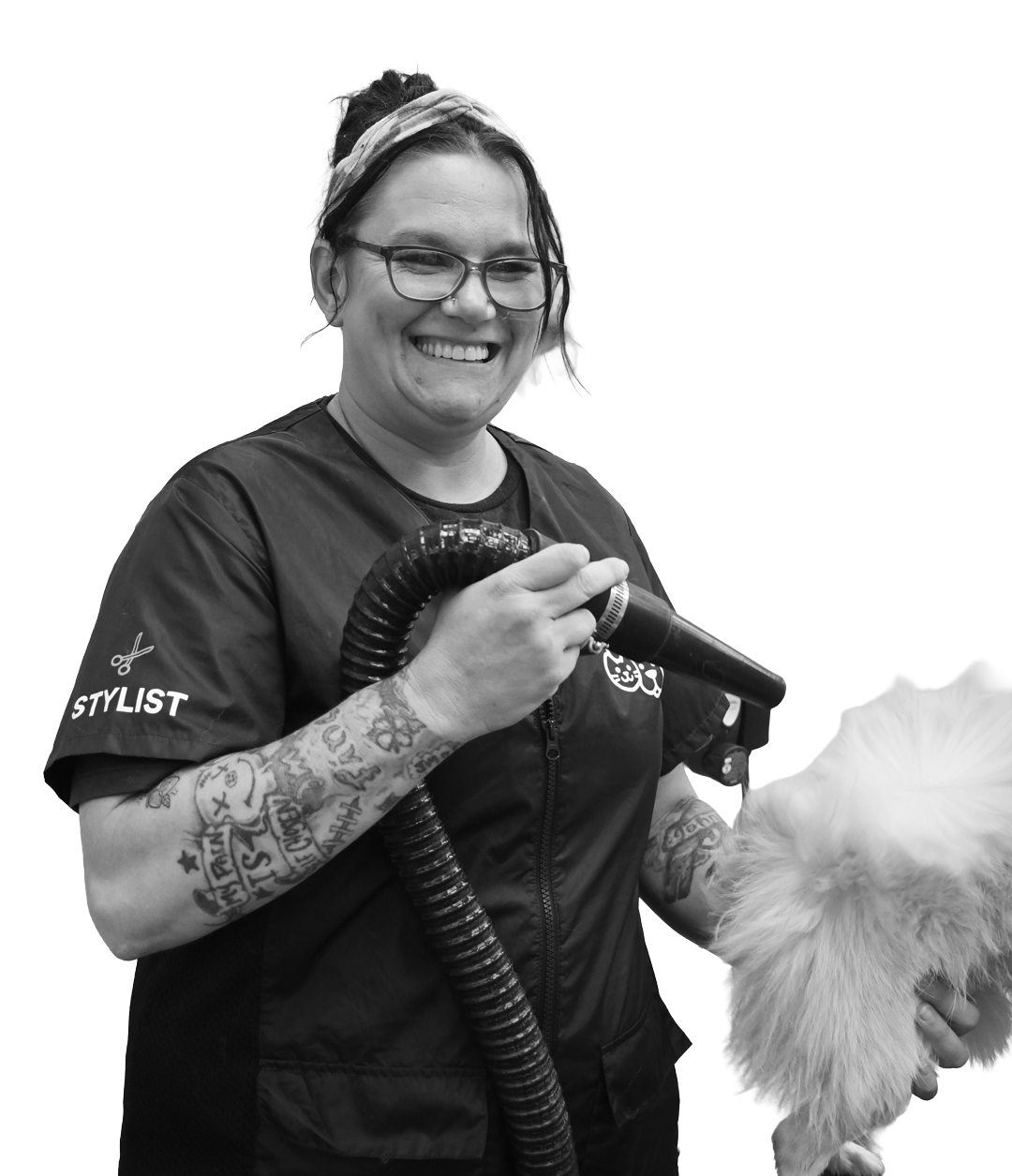A smiling pet groomer with tattoos uses a drying tool on a fluffy dog. The groomer, featured in the press kit, wears glasses and a uniform labeled "STYLIST.