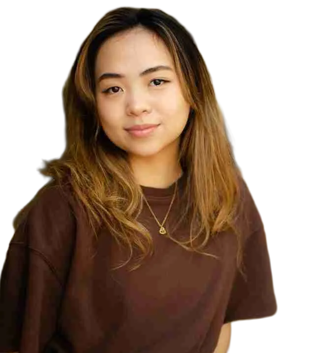 A young woman with long, light brown hair wearing a brown shirt and blue pants sits and looks at the camera against a plain background, perfect for a press kit.