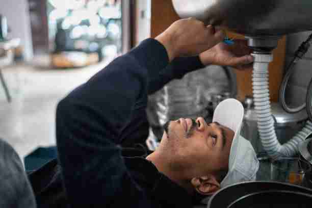 A person wearing a cap fixes plumbing under a kitchen sink, using a tool to work on the pipes with the precision of an Administrative Medical Assistant handling important tasks.