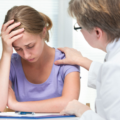 A doctor or CNA places a reassuring hand on the shoulder of a distressed woman who is sitting at a desk, holding her head in her hand.
