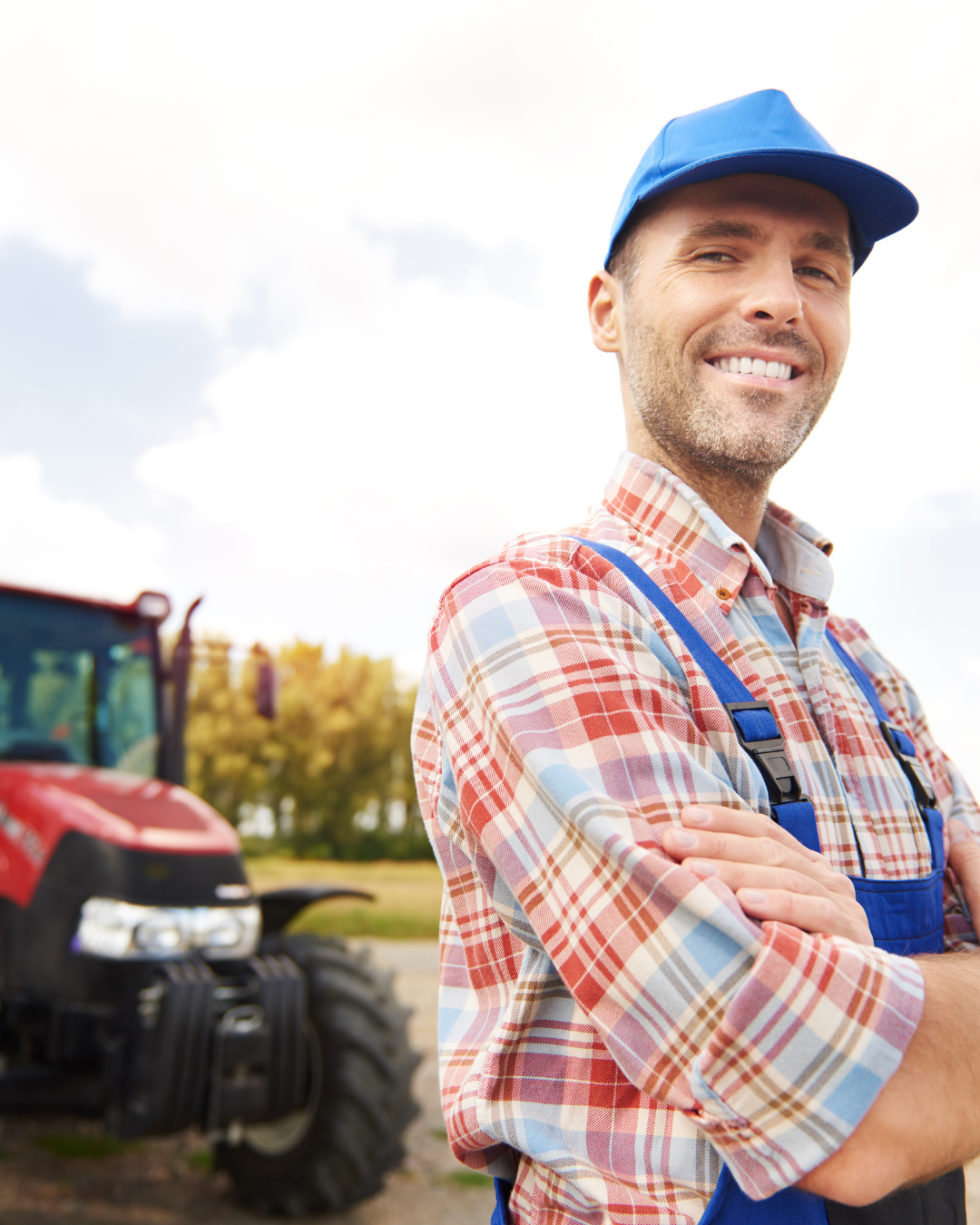 Man in plaid shirt and blue cap stands with crossed arms, smiling in front of a red tractor outdoors—a proud representation of agriculture careers.