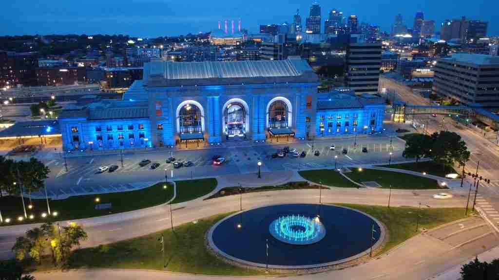 A large historic train station building is lit with blue lights at dusk, with a circular fountain in front and the Kansas City Celebrates Our Scholars city skyline glowing in the background.