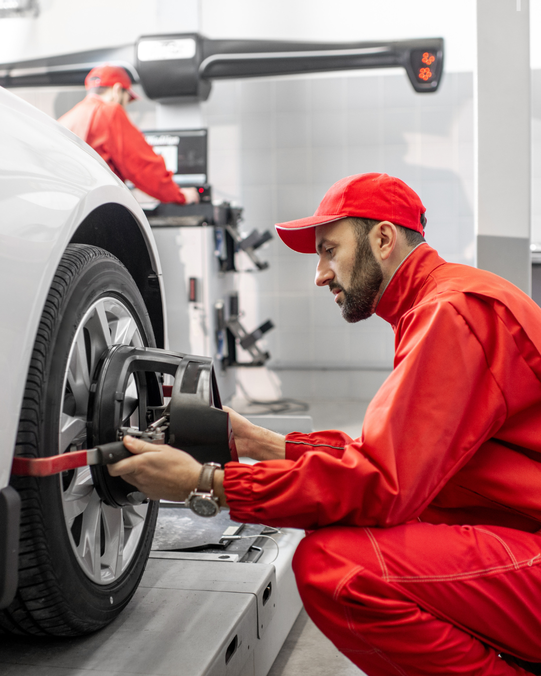 An automotive technician in a red uniform adjusts equipment on a car wheel in a workshop, while another technician works in the background.