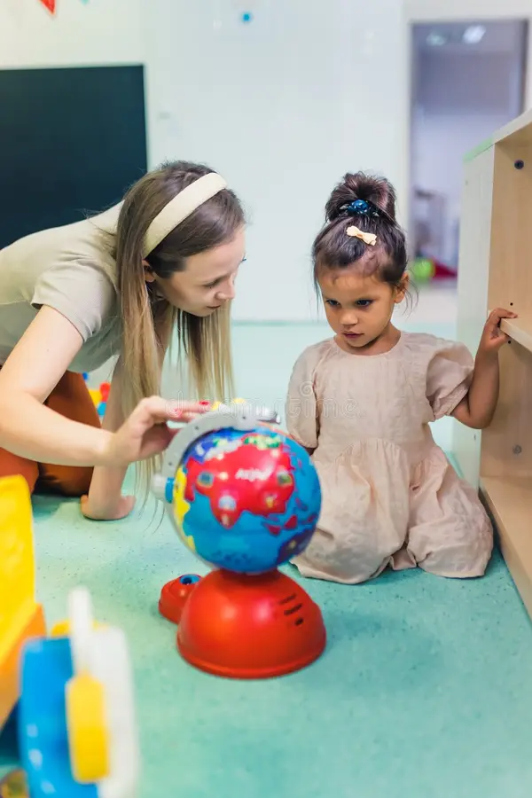 An Early Childhood Educator and a child are kneeling on the floor, looking at a toy globe together in a classroom setting.