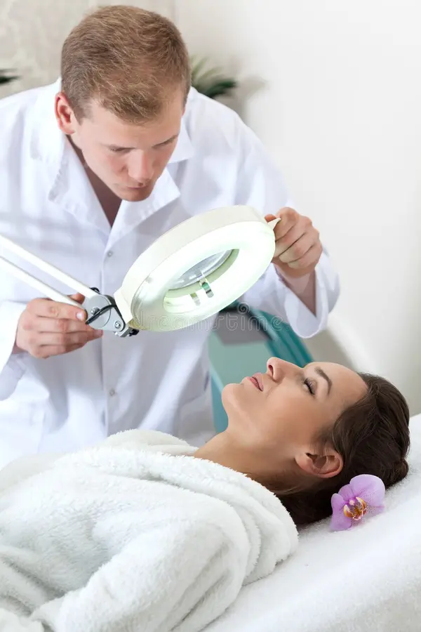A dermatologist explores a woman's face with a magnifying lamp as she lies on a treatment table, wearing a white robe with a flower in her hair—showcasing the expertise found in cosmetology careers.