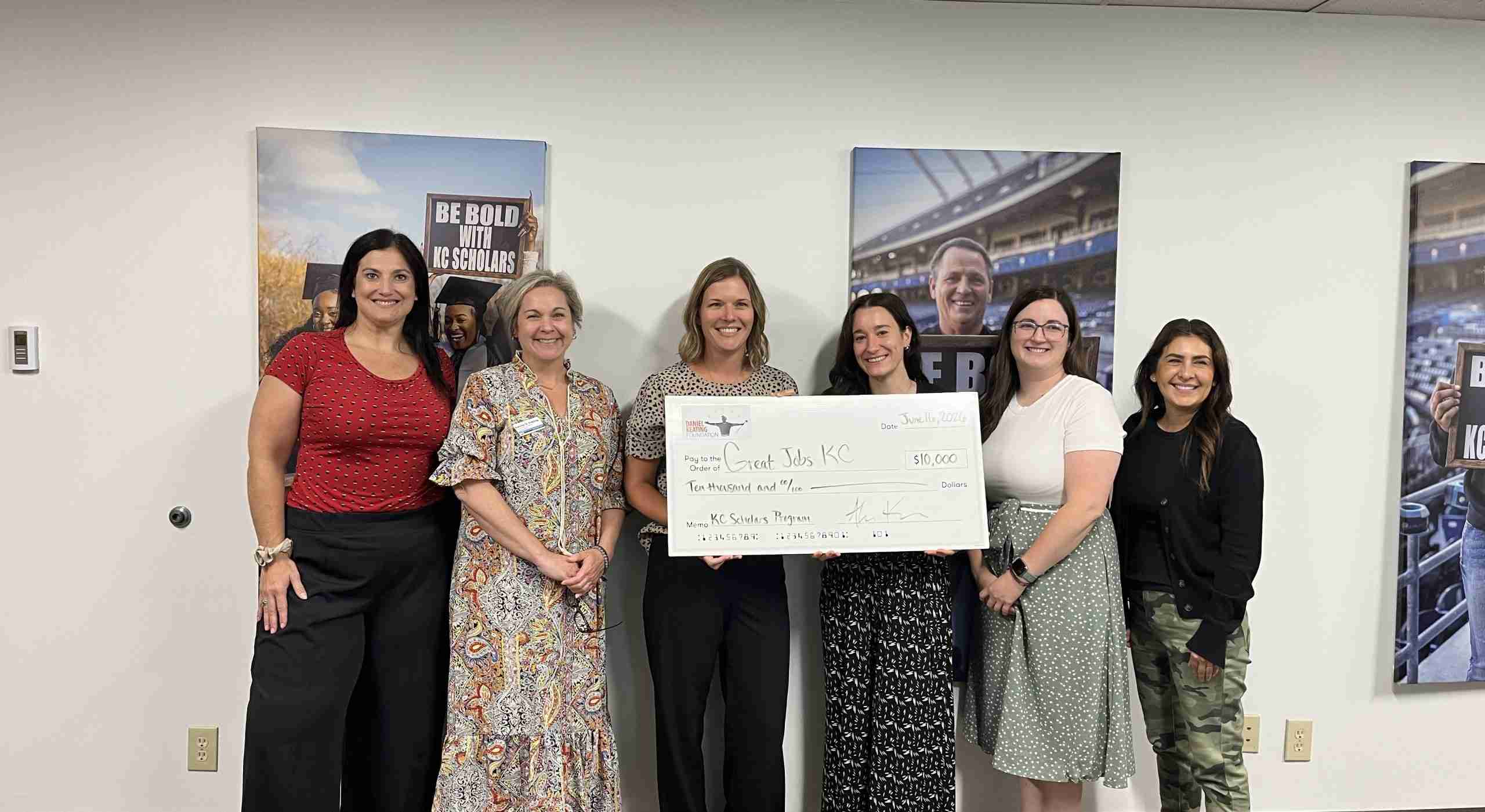 Six women stand indoors, smiling and holding a large check for $10,000 addressed to Great Jobs KC. Motivational posters and "The world is watching KC" slogan are visible on the wall behind them.