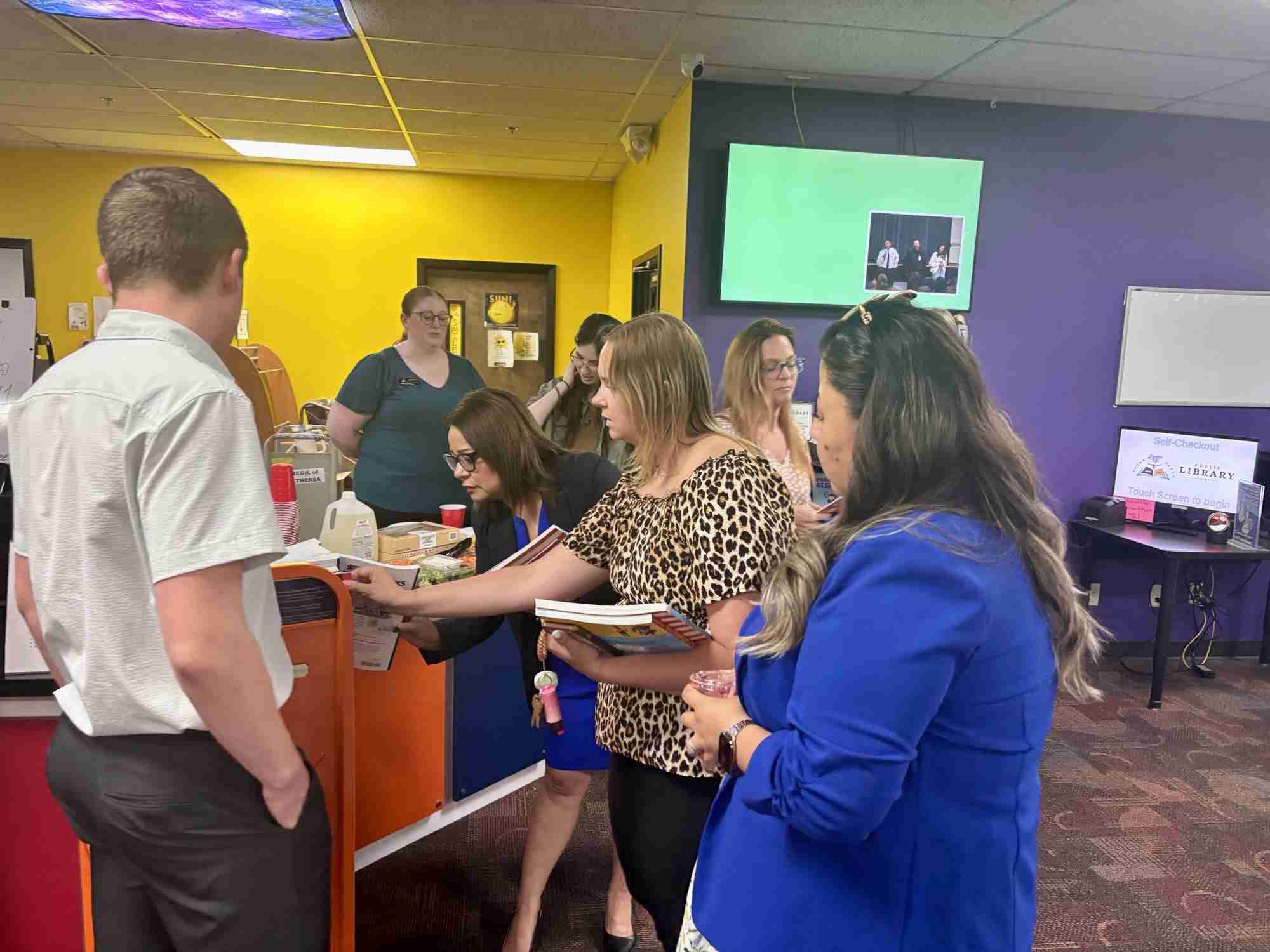 A group of adults stand and interact near a colorful counter in a library or office space, with books and papers in hand—gathered to celebrate Ten Years of Scholars, marking Ten Years of Impact.