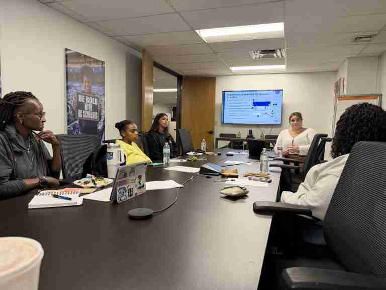 Five people sit around a conference table, listening to a woman presenting slides on a screen about college scholarships and skills training. Laptops, notebooks, and water bottles are visible on the table, adding to the focused atmosphere as participants prepare for the back to school season.