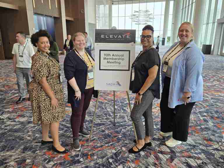 Four women stand beside a sign for the 90th Annual Membership Meeting in a conference hall, where other attendees gather to discuss important topics like back to school initiatives, college scholarships, and skills training programs visible in the background.